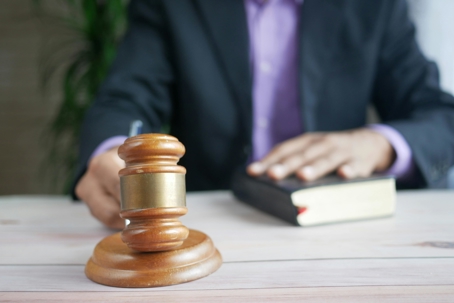 Judge’s gavel on a desk in the foreground, with a person in a suit placing a hand on a book in the background, suggesting a courtroom or legal setting.